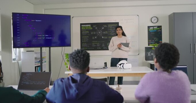 Diverse class teacher using tablet teaching code while monitor showing, teens typing laptops VR set