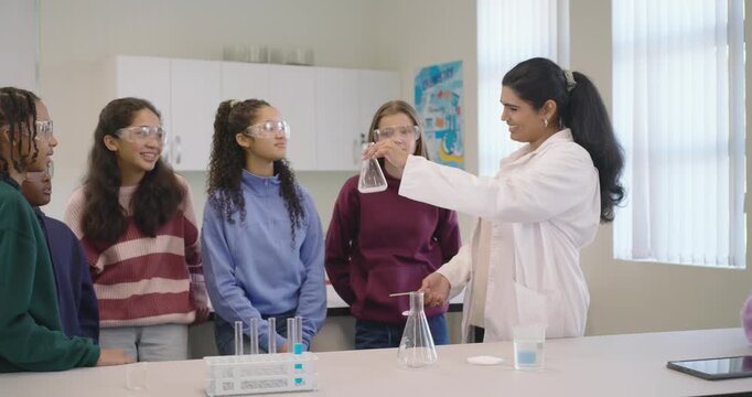 Indian instructor holding flask at lab bench, rotating, offering while students reaching, learning