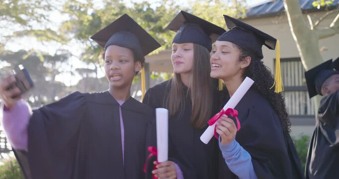 Diverse female graduates in gowns huddling around phone taking selfies on campus holding diplomas