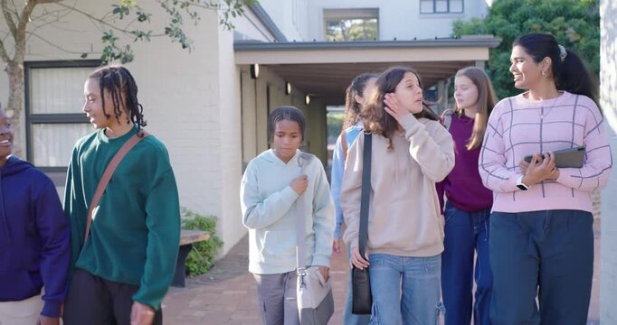 Diverse teens and mentor in pink sweater exiting path walking into quad bags tablet between classes