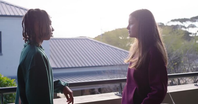 Diverse teenage friends talking after left leaning on balcony railing, smartphone and bag strap