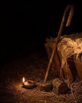Wooden shepherd crook leaning against rustic wooden manger on straw in stable. Nativity scene with oil lamp and vintage treasure chest. Biblical background for Christmas holiday concept.