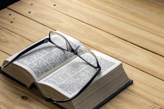 Black framed reading eyeglasses resting on open dictionary book with visible printed word definitions on a rustic wooden plank table, concept of nearsightedness, education, learning and study