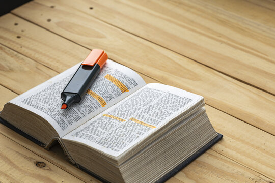 Open thick English dictionary book with orange highlighter marker pen resting on pages showing highlighted vocabulary words on rustic wooden table surface for learning and language education concept
