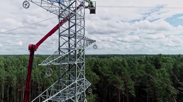 Latvia - 13 February 2023: Aerial view of a technician working on a high-voltage electricity pylon with a crane nearby, surrounded by a dense forest.