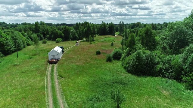 Latvia - 19 February 2023: Aerial view of a truck pulling a white modular structure along a dirt road through a lush green forest landscape under a cloudy sky.