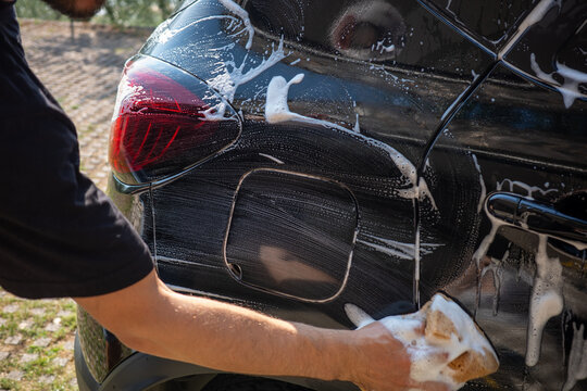 Person washing a car with soap and a sponge in a sunny outdoor area