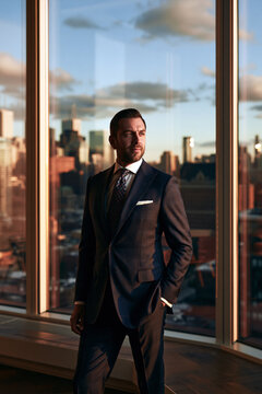 Full-length portrait of a confident lawyer standing in a luxury office, wearing an elegant tailored suit with a city skyline visible through tall windows.