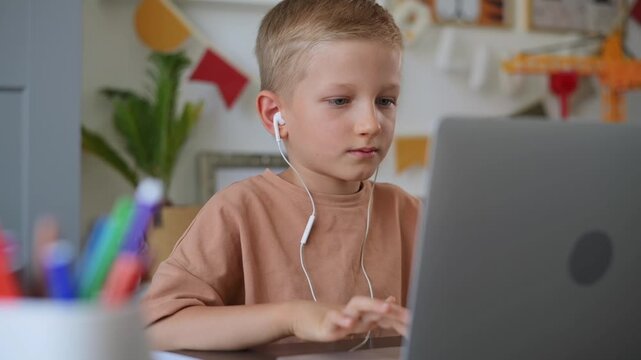 Cute boy wearing headphones works diligently on laptop. Modern technology: child surfs the internet, plays computer games, watches an online lesson, and video chats with teacher. Slow motion