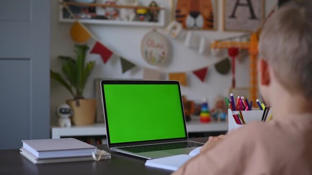 Schoolboy using laptop with green screen for online lesson at home. Computer display supports chroma key replacement for education, digital learning, remote classes, creative media production