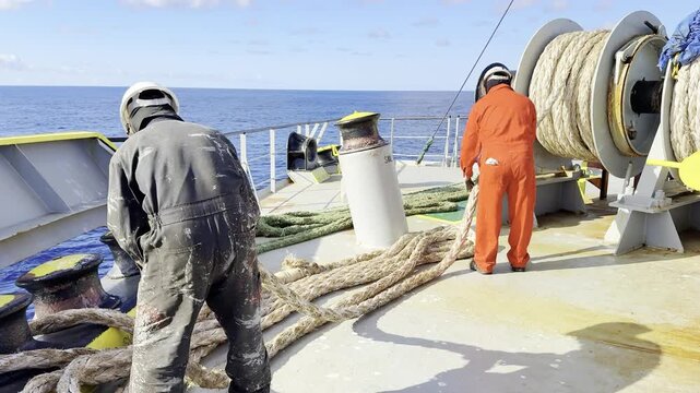 Ship crew uncoiling heavy ropes from winches at sea.