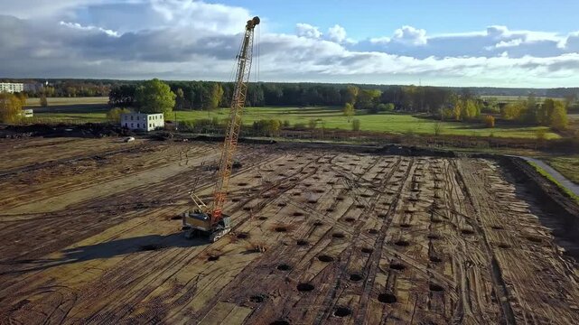 Latvia - 18 February 2023: Aerial view of a construction site featuring a large yellow crane and rows of drilled holes in the dirt field under a cloudy sky.