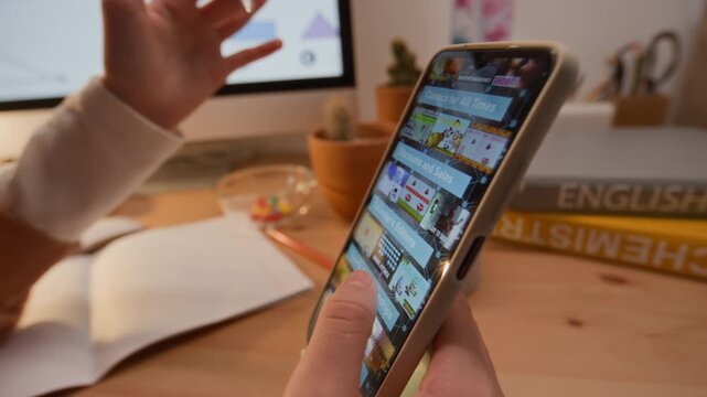 Unrecognizable teenager scrolling on smartphone while studying at desk with textbooks and computer. Footage is perfect for educational marketing, digital distraction concepts, or student lifestyle ads