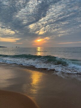 A glowing golden reflection stretches across wet sand as a transparent Atlantic wave gently curls toward the shore. Dramatic textured clouds glow beneath the warm sunset sky.