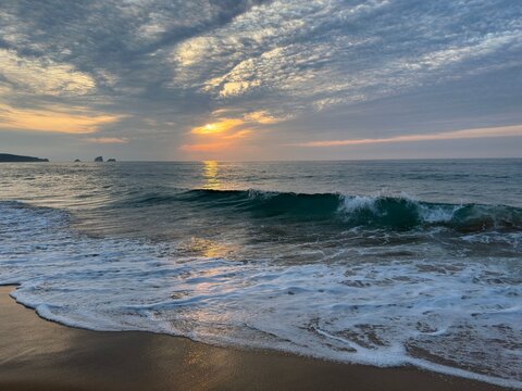 A rising dark emerald wave catches the last rays of sunset over the calm Atlantic. Dramatic textured clouds glow above shimmering reflections, creating a peaceful sunset with natural color and light.