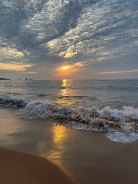 A breaking Atlantic wave bursts into white sea foam beneath a glowing sunset.Golden reflections shimmer across wet sand while dramatic textured clouds fill the evening sky with natural color and light