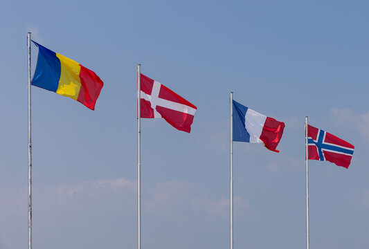 Romania, Denmark, France and Norway flags form a descending row of four poles, waving in daylight above a soft cloud band.