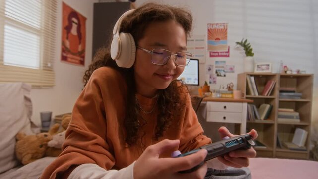 Asian teenager wearing headphones and glasses playing handheld console in bedroom. Footage represents youth entertainment, gaming technology, and modern leisure activities