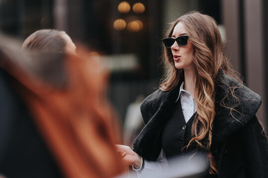 A young professional woman wearing sunglasses and a coat speaks while gesturing outdoors. She appears engaged in a city business conversation with a colleague.