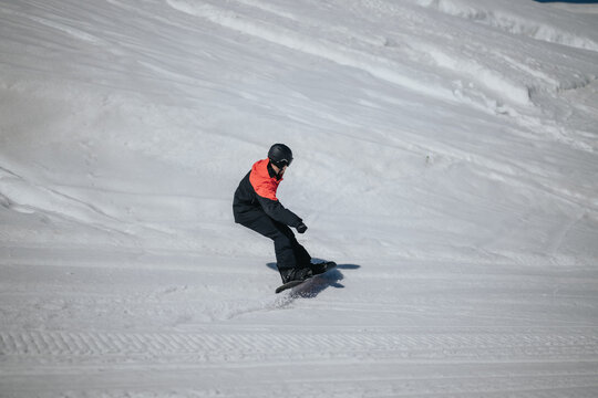 A snowboarder in a red and black jacket carves down a groomed snowy slope. The winter scene shows controlled snowboarding on a sunlit mountain run.