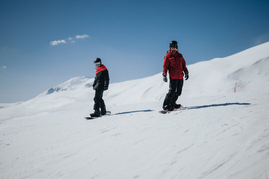 Two snowboarders in red jackets ride down a snowy mountain slope wearing helmets. A bright winter scene shows them snowboarding side by side under a clear blue sky.