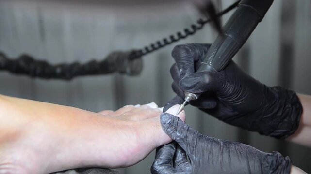 Close-up of a professional nail technician using an electric nail drill with a ceramic bit to remove old gel polish from a client's toenail during a pedicure. Professional foot care.