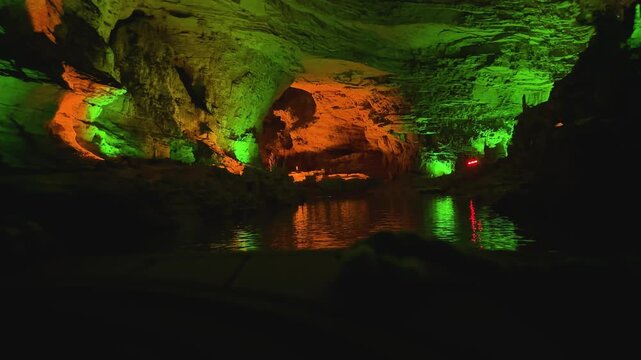 Illuminated underground cave with river reflection. Green orange rock formations inside natural grotto. Travel destination for cave exploration tour adventure. Tourists traveling by boat time lapse.