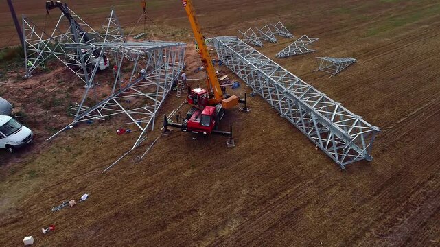Latvia - 18 February 2023: Aerial view of a yellow crane installing a large metal power line pylon in an open field with workers on site.