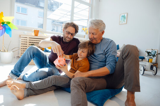 Senior man with son and grandson putting money in piggy bank at home
