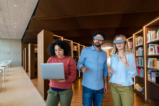 Man and woman with VR glasses gesturing by colleague using laptop in library