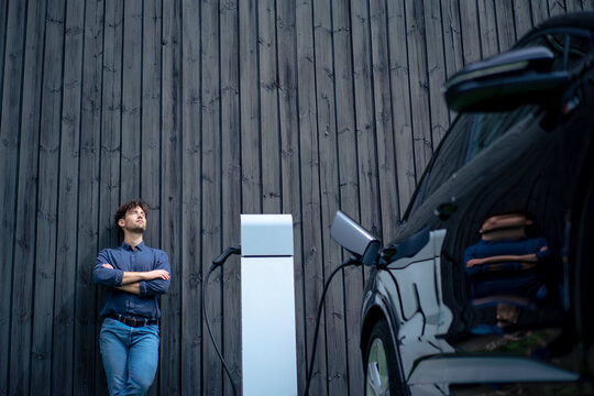 Man with arms crossed leaning on wall by electric car