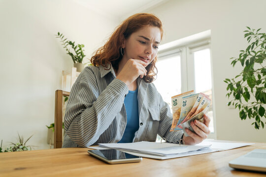 Confused young woman calculating financial bills and counting currency at home