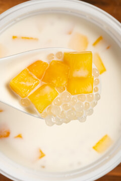 Spoon filled with mango cubes and sago above a bowl of creamy mango sago dessert