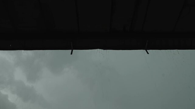 A low-angle shot of rain pouring from a dark roof gutter against a gloomy, overcast sky.