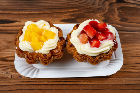 Two flaky puff pastry tarts topped with cream, fresh mango cubes and sliced strawberries on a white paper plate