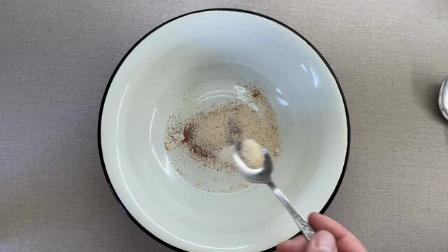 Hand adding garlic powder and paprika to white bowl on neutral background