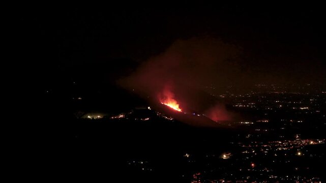 A massive wildfire burns fiercely on a dark mountain at night above the glowing city lights of Sermoneta. Such devastating natural disasters highlight the urgent global crisis of climate change.