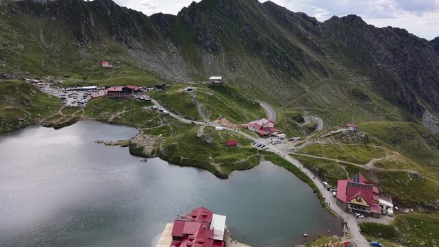 Forward and ascending drone flight revealing B&acirc;lea Lake, mountain restaurants, visitors and parked vehicles surrounded by the dramatic peaks of the Făgăraș Mountains in Romania.
