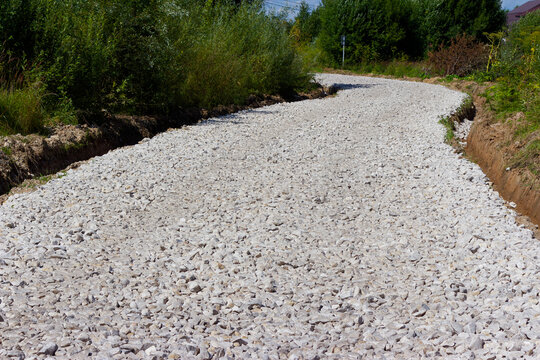 Newly laid gravel road winding through lush green summer countryside, crushed stone path under bright daylight, industrial road construction materials outdoors