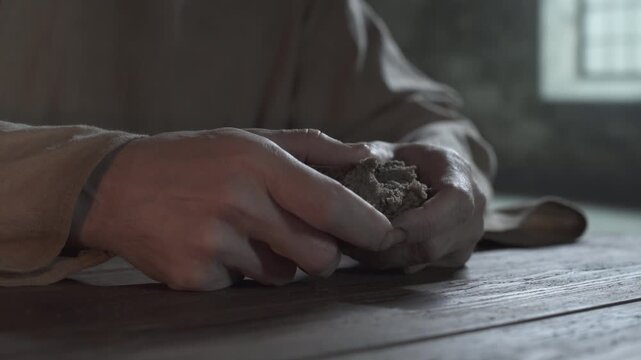 Hands of a medieval prisoner slowly breaking a piece of bread on a rustic wooden table inside a gloomy dungeon