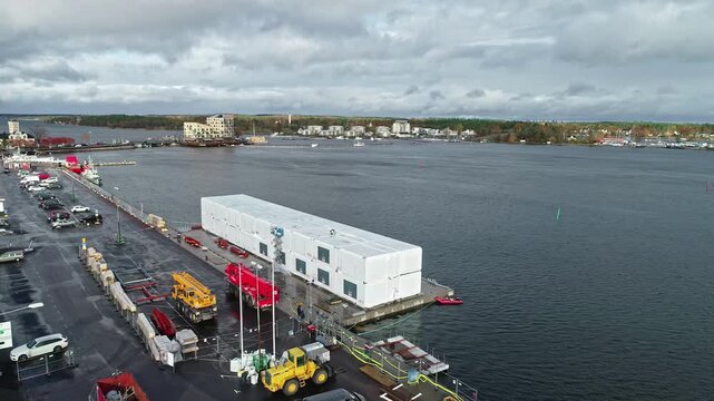 Sweden - 19 February 2023: Aerial view of a harbor construction site featuring a large white modular building, yellow construction cranes, and vehicles parked along the waterfront.
