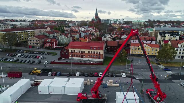 Sweden - 19 February 2023: Aerial view of a construction site with red mobile cranes lifting a white modular unit in a Swedish town with historic buildings and a church spire.