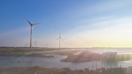 Fototapeta premium Wind turbines spinning over coastal wetlands in Yuanli, Miaoli, Taiwan during a hazy golden sunset with beautiful light flares.