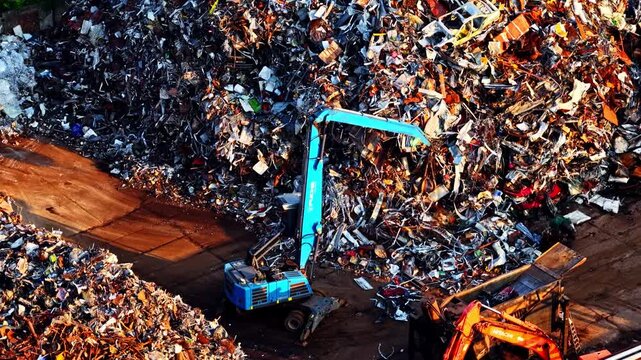 Riga, Latvia - 26 August 2025: Aerial view of an industrial scrapyard with a blue excavator sorting through large piles of scrap metal and discarded vehicles.