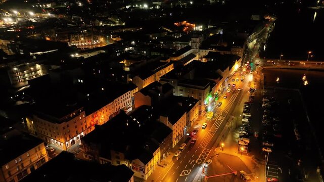 Cinematic drone view of festive Waterford beside the River Suir, with illuminated buildings, Christmas decorations, glowing streets, and moving traffic after dark