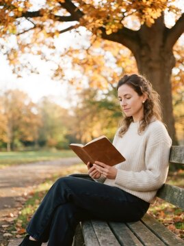 Woman sitting on bench in park reading book. Female reader enjoying literature outdoors during autumn season. Leisure activity and lifestyle concept in nature with golden fall foliage.