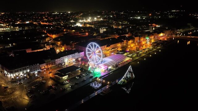 Cinematic aerial reveal of Waterford after dark with an enormous Christmas Ferris wheel dominating the skyline above the illuminated riverfront and busy streets.