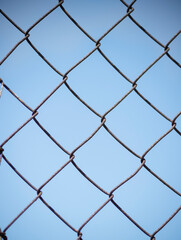 A metallic chain link mesh fence stands against a clear blue sky background. The steel wire grid creates a repeating diamond geometric pattern. © dsbedarieva