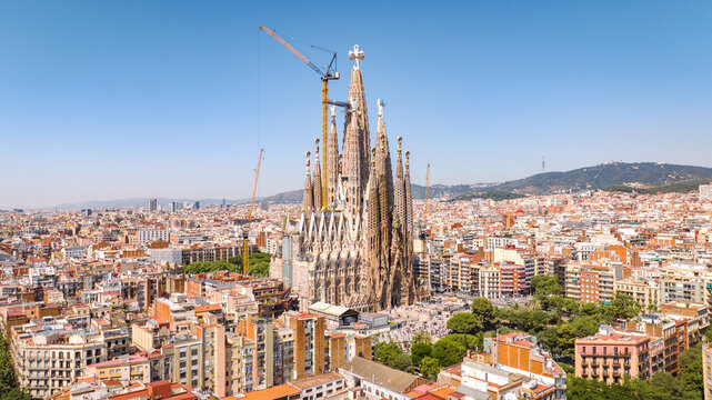 Panoramic drone view of Barcelona with the iconic Sagrada Familia, dense urban skyline, green boulevards and Catalan hills on a sunny summer day.