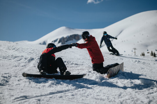 Two snowboarders on a snowy mountain slope help one another while kneeling and holding hands. A third snowboarder rides past in the background down the sunlit run.